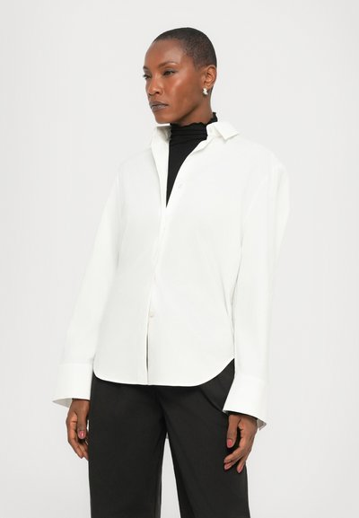 Chemise blanche à manches longues avec col, en tissu lisse et coupe décontractée, portée par-dessus un haut noir. Le modèle pose avec les mains détendues sur les côtés.