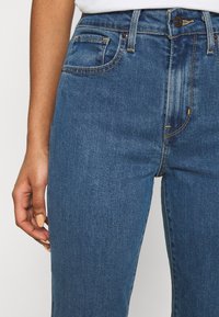Jeans de mezclilla azul con un diseño recto, que cuentan con un cierre frontal de botón, cinco bolsillos y detalles de costura en contraste.