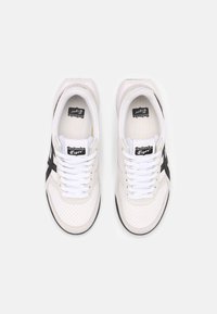 White athletic shoes with black accents and perforated design. Features flat laces and a cushioned sole. Branding visible on the tongue.