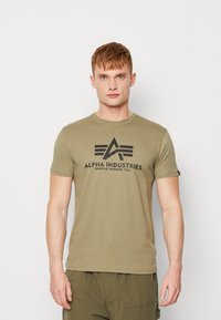 Olive zaļa kokvilnas t-krekls ar apaļu kakla izgriezumu, īsām piedurknēm un melnu grafiku, kas attēlo tekstu "ALPHA INDUSTRIES."