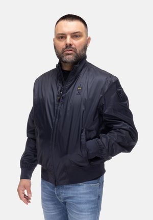 Uomo con barba che indossa una giacca bomber blu navy con zip e jeans azzurri, in piedi con una mano in tasca davanti a uno sfondo bianco.