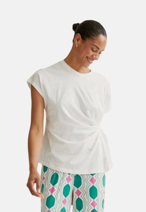 Mujer con el pelo recogido en un moño, lleva una camiseta blanca de manga corta con detalle de torsión lateral y pantalones estampados con formas en verde azulado y rosa.