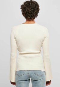 Ribbad krämfärgad långärmad topp med åtsittande midja och breda ärmar, i kombination med ljusblå jeans. Slät textur och subtila designdetaljer.