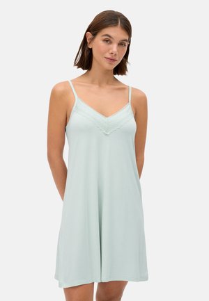 Mujer con camisón sin mangas azul claro con detalles de encaje en el escote en V, de pie contra un fondo liso.