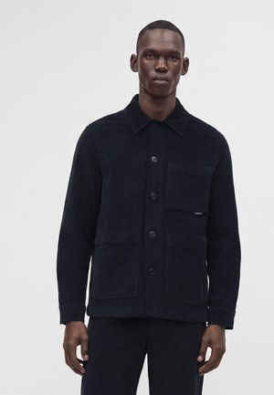 Homme portant une veste noire boutonnée avec trois poches avant sur un fond uni gris clair.