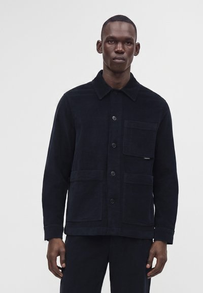 Homme portant une veste noire boutonnée avec trois poches avant sur un fond uni gris clair.