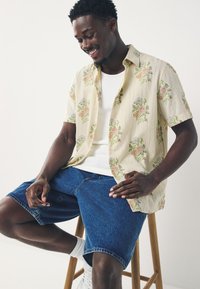 Camicia crema a maniche corte con motivo floreale, abbinata a pantaloncini di jeans blu. La modella è seduta su uno sgabello di legno, indossa calzini bianchi e sneakers.