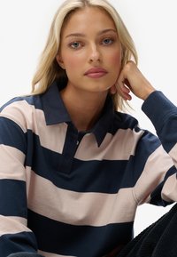 Blonde vrouw met blauwe ogen die haar hoofd op haar hand steunt, gekleed in een marineblauw en lichtroze gestreepte rugbyshirt tegen een witte achtergrond.