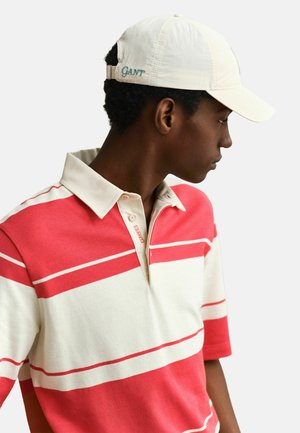Junger Mann, der ein rot-weiß gestreiftes Poloshirt und eine weiße Kappe mit der seitlichen "GANT"-Stickerei trägt, schaut nach rechts.