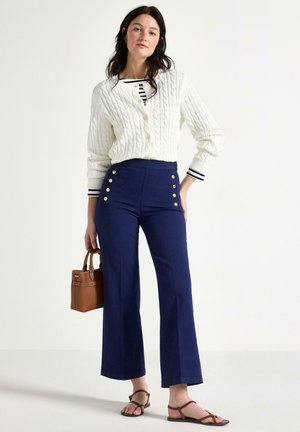 Donna che indossa un cardigan bianco a trecce sopra una camicia a righe, pantaloni a vita alta blu navy con bottoni dorati, tiene in mano una borsa marrone, in piedi a piedi nudi.