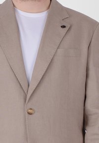 Blazer di lino beige con chiusura a un bottone, rever a sbieco e taschino sul petto. Indossato sopra una maglietta bianca semplice.