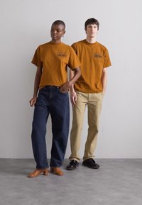 Dwa modele w dopasowanych brązowych koszulkach z logo "Aries", zestawionych z luźnymi niebieskimi dżinsami i beżowymi spodniami, stojący na szarym podłożu.
