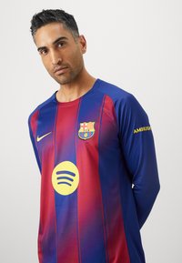 Dugine dugih rukava Barcelona FC u crvenoj i plavoj boji s vertikalnim prugama. Sadrži Nike logo, grb kluba i žuti krug dizajna Spotify.