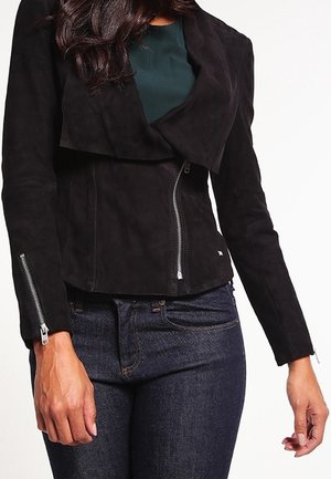 Veste en suède noir avec un devant drapé, fermeture éclair et manches longues. Portée par-dessus un haut sombre et assortie à un jean en denim.