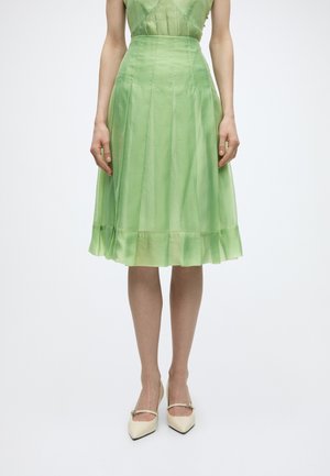 Kobieta w jasnzielonej plisowanej spódnicy do kolan i kremowych szpilkach z paskami wokół kostki, stojąca na jednolitym tle.