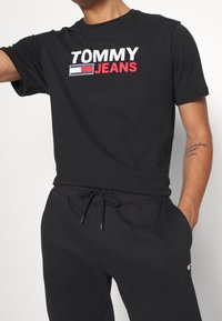 Svart bomullströja med korta ärmar och en iögonfallande vit och röd "TOMMY JEANS" logotyp. Matchas med svarta joggingbyxor med elastisk midja.
