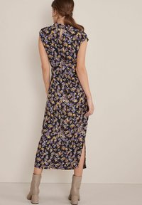 Kobieta stojąca tyłem, ubrana w czarną midi sukienkę w kwiaty w fioletowe i żółte wzory oraz beżowe botki sięgające kostek.