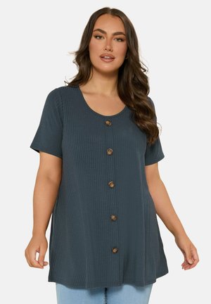 Kvinde med langt, bølget hår iført en mørkeblå ribbet kortærmet tunika med fem brune knapper foran, kombineret med lyseblå jeans.