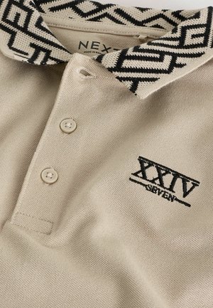 Beige polo shirt with a patterned black and white collar, three buttons, and "XXIV SEVEN" embroidered in black on the front. Cotton material.