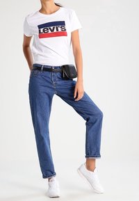 Camiseta blanca con un logo de Levi's en rojo y azul marino, combinada con jeans azules y zapatillas blancas. Riñonera negra llevada en la cintura.