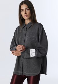 Camicia grigia a bottoni realizzata in un morbido tessuto a maglia, con taschino frontale, cuciture in contrasto e etichetta sul polsino. Le aperture laterali aggiungono un tocco di dettaglio.