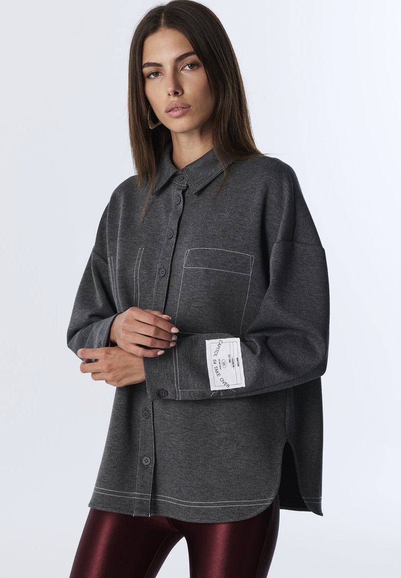 Camicia grigia a bottoni realizzata in un morbido tessuto a maglia, con taschino frontale, cuciture in contrasto e etichetta sul polsino. Le aperture laterali aggiungono un tocco di dettaglio.