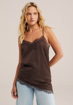 Blondinka, oblečena v temno rjavo kamisolko z čipko in svetlo modre kavbojke, rahlo obrnjena v stran, na enostavni podlagi.