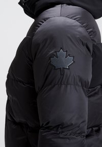 Veste en duvet noire avec un emblème texturé en forme de feuille d'érable sur la manche gauche, finition lisse et brillante et design matelassé.