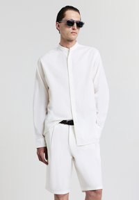 Camicia bianca a maniche lunghe con colletto alto, tessuto testurizzato, abbinata a pantaloni corti bianchi e una cintura intrecciata scura. Occhiali da sole neri completano il look.
