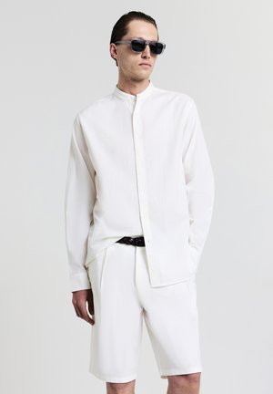 Chemise blanche à manches longues avec un col montant, en tissu texturé, associée à un short blanc et une ceinture tressée noire. Des lunettes de soleil noires complètent le look.