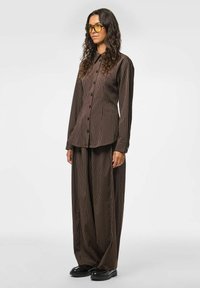 Ruda dryžuota marškinėlių ir plačios kelnių komplektas, pasižymintis sagų užsegimu, ilgomis rankovėmis ir laisvu siluetu. Komplektą sudaro juodi batai ir geltoni akiniai.