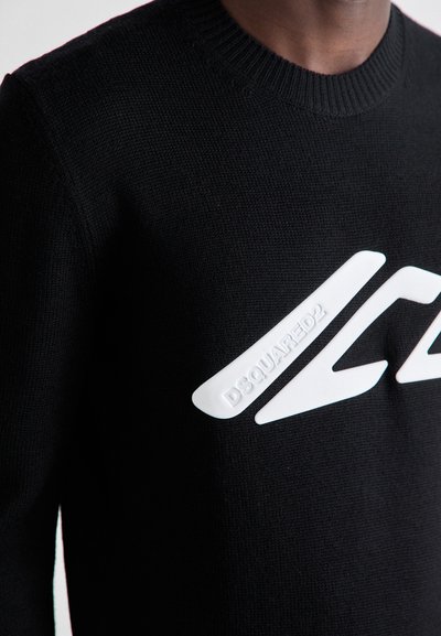 Sort striktrøje med ribbet rund hals og en tekstureret hvid logo detalje foran. Tæt på udsigt af stoffet og designet.
