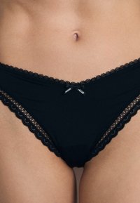 Culotte en dentelle noire avec des bords festonnés, fabriquée en tissu doux, avec un branding subtil et un petit nœud à l'avant.