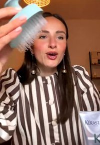 Frau mit langen dunklen Haaren und gestreiftem Shirt hält einen blauen Kopfhautmassager und ein Kérastase Haarprodukt auf einem Tisch.