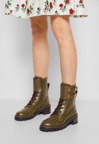 Stivaletti militari verde oliva con finitura lucida, lacci, cerniere, fibbie dorate e una suola in gomma nera spessa, indossati con un vestito floreale.