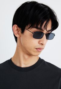 Rechteckige schwarze Sonnenbrille mit dünnen Drahtgestellen. Getragen von einer Person in einem schwarzen T-Shirt, vor einem schlichten hellen Hintergrund.