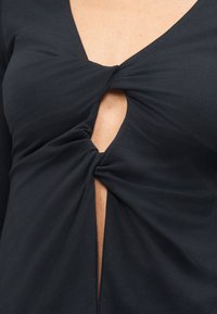 Μαύρο τοπ με V λαιμόκοψη και δύο στριφογυριστούς κόμπους με ανοίγματα που αποκαλύπτουν δέρμα στην άνω περιοχή του στήθους και στη μέση του κορμού.