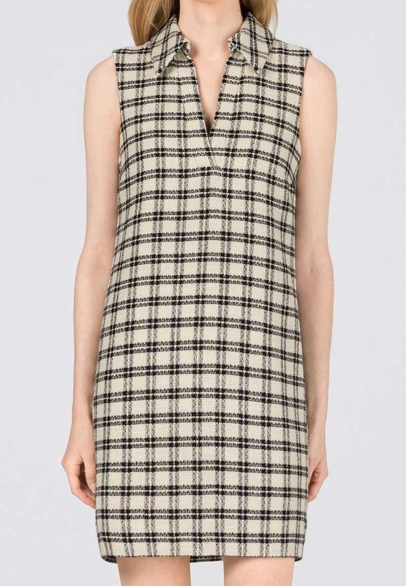 Femme portant une robe crème sans manches, longueur genou, à motif à carreaux noirs et large col, debout sur un fond uni.