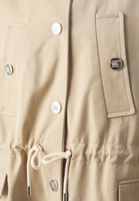 Chaqueta beige con botones a presión plateados, cinturilla con cordón y dos bolsillos frontales con bordes cosidos. Textura de tela de algodón suave.