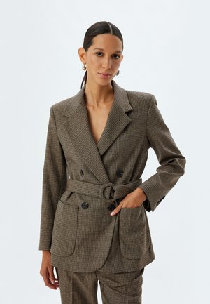 Blazer a quadri marroni con bottoni doppi, ampie rever e vita cintura; presenta tasche a toppa e una texture del tessuto liscia.