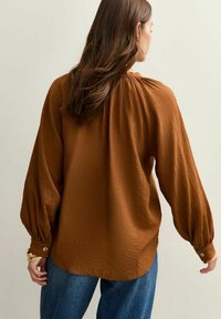 Blouse marron à texture avec des manches bouffantes, accents de boutons aux poignets, portée sur un denim bleu clair, vue de l'arrière.
