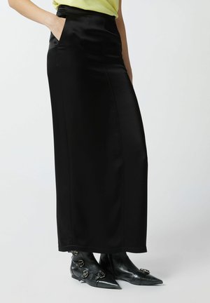 Maxi gonna nera in satin con tasche laterali, caratterizzata da una texture liscia e linee pulite, abbinata a stivaletti neri a punta.