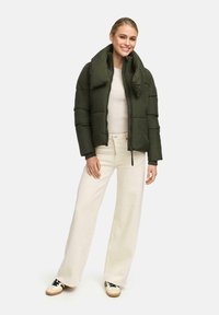 Young woman smiling, wearing an olive green puffer jacket over a beige top, cream wide-leg pants, and white sneakers with black laces.