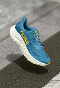 Zeleno-modra tekaška obutev z rumenimi poudarki, mrežasti zgornji del, oblazinjen podplat in sistema za zavezovanje. Na strani je jasno prikazana blagovna znamka.