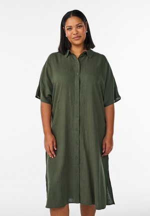 Žena s čiernymi vlasmi dĺžky po ramená, oblečená v voľných, zapínateľných olivovo zelených šatách pod kolená na bielom pozadí.