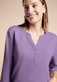 Maglietta a maniche lunghe viola con scollo henley, quattro bottoni e una texture morbida. Presenta una vestibilità comoda e un design semplice.