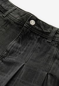 Sorte denimbukser med plisseret frontdesign, udstyret med en sølvfarvet knaplukning og tekstureret stof, der fremhæver syningerne.
