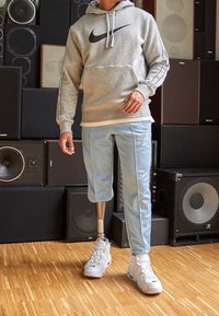 Grå Nike-hettejacka med svart logotyp, ljusblå Nike-svettdresser och vita sneakers. Innehåller ett prostetiskt ben. Bakgrunden har högtalare.