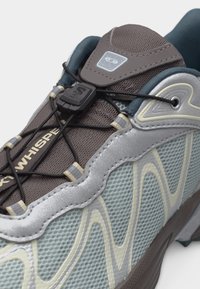 Približek sive in bež tekaške copate z mrežastim tekstilom, oblikovanimi valovitimi vzorci in hitrim črnim vezalkam, pritrjenim s plastičnim zaponkom.