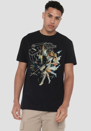 Junger Mann trägt ein schwarzes Star Wars Grafik-T-Shirt und beige Cargo-Shorts und steht vor einem einfarbigen Hintergrund.
